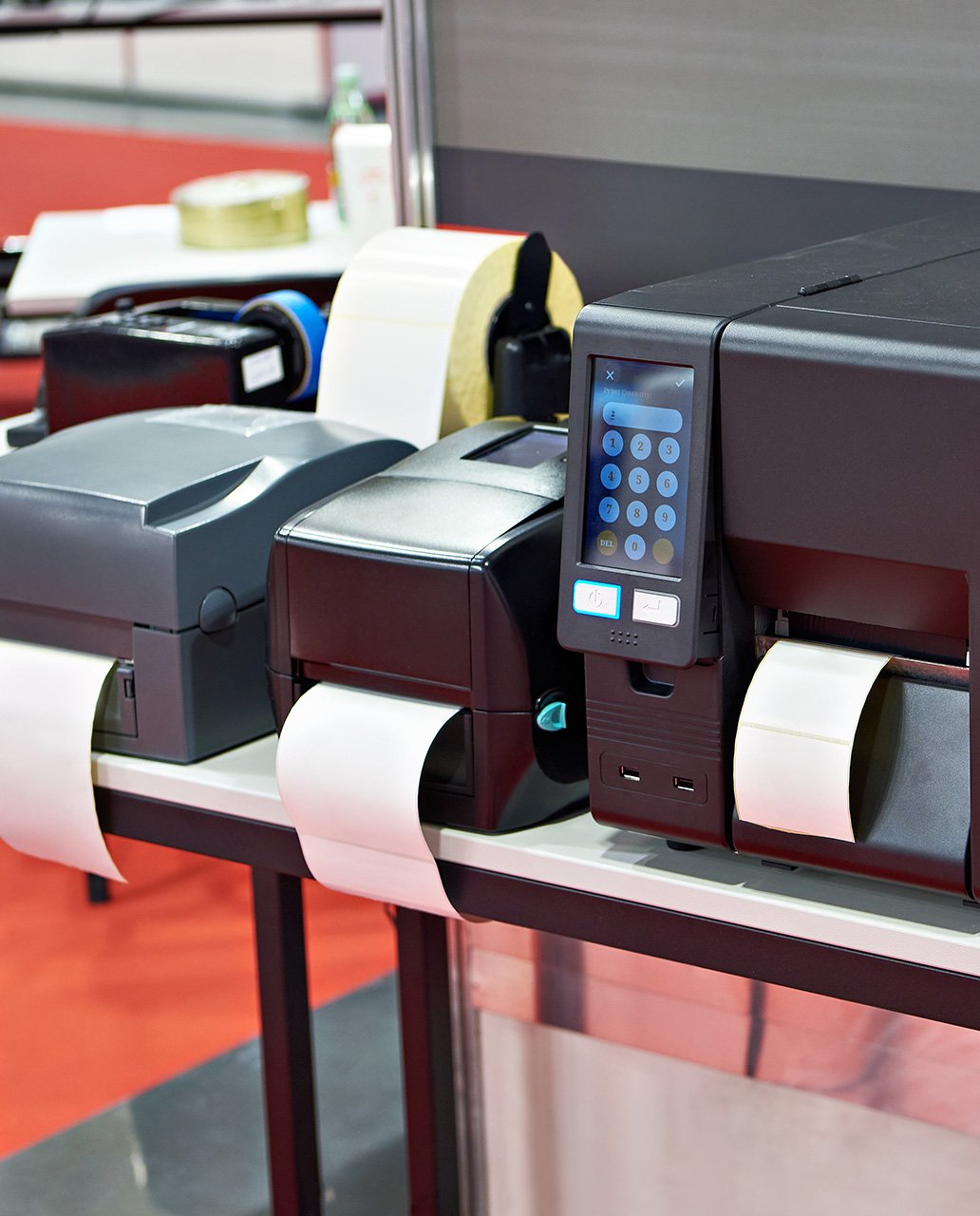 Gros plan de plusieurs imprimantes d'étiquettes sur une table, imprimant des étiquettes sur du papier blanc, l'une d'entre elles affichant un panneau de commande numérique.